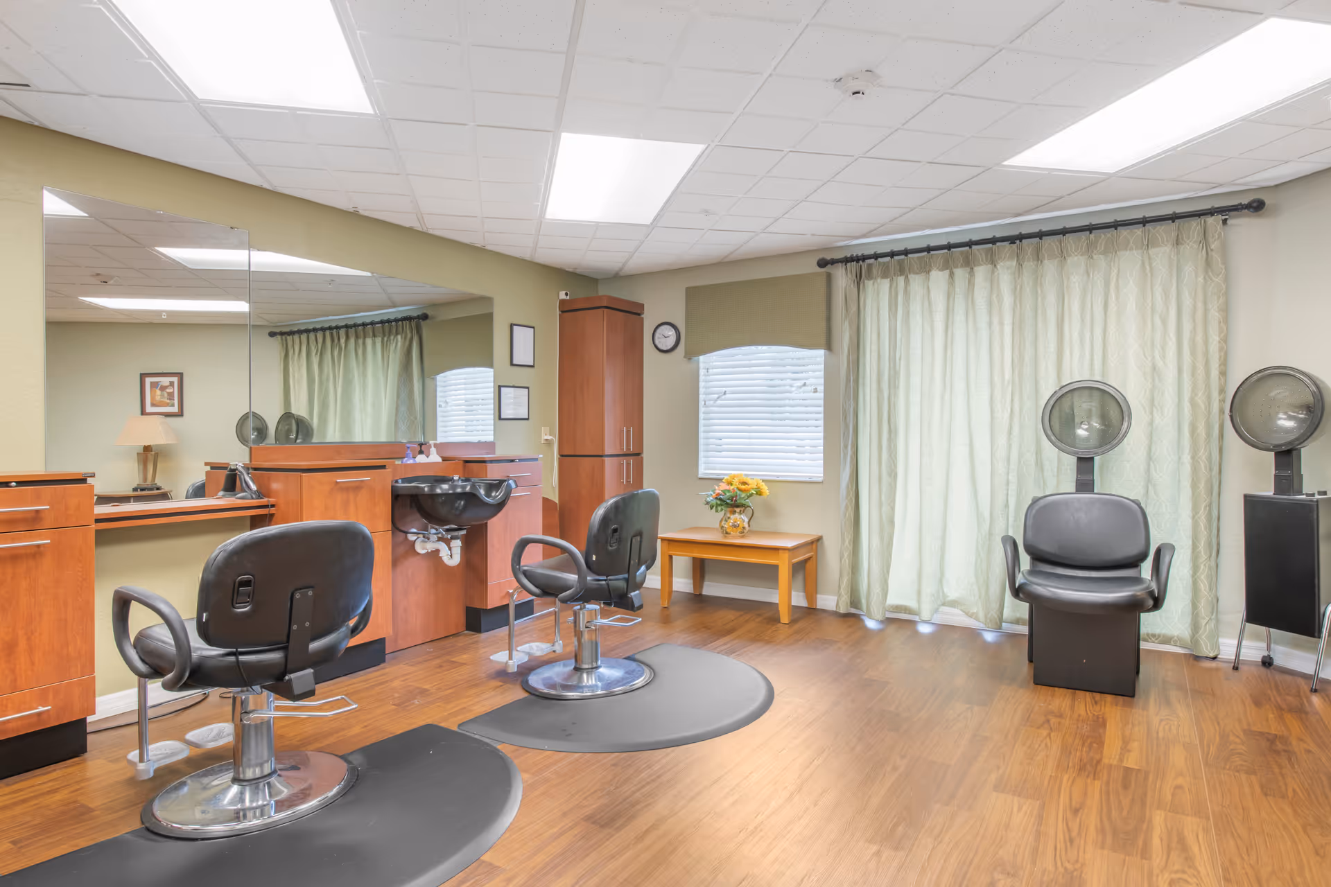 Bright salon room with styling chairs, a wash sink, hooded hair dryers, mirrors, and wooden cabinetry.