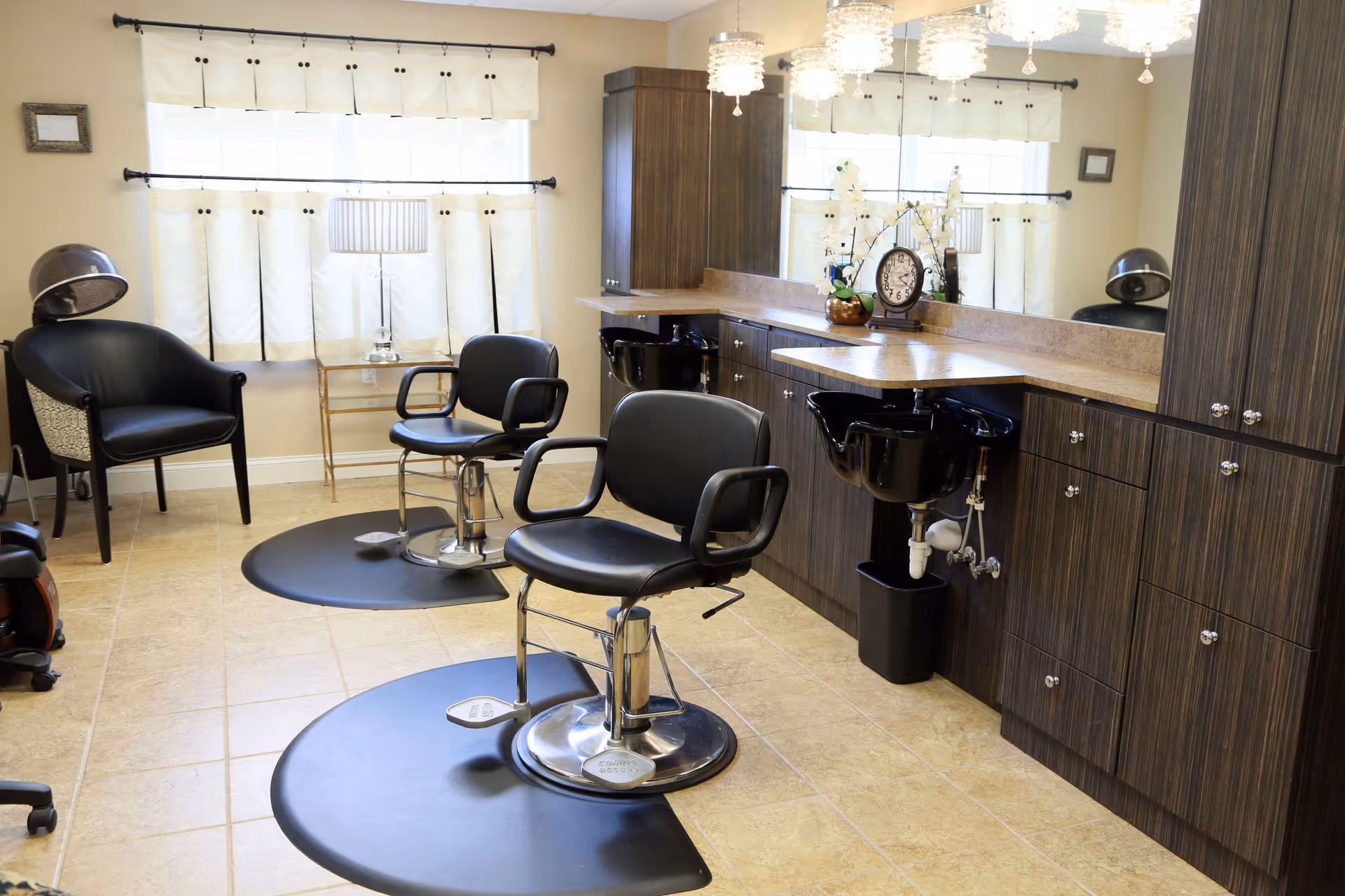 Interior of a salon area with two black salon chairs on black mats in front of a counter with sinks and dark wood cabinets. There are two large windows with white curtains, a small table with a lamp, and a black armchair in the corner. The floor is tiled and the room is well-lit with ceiling lights.