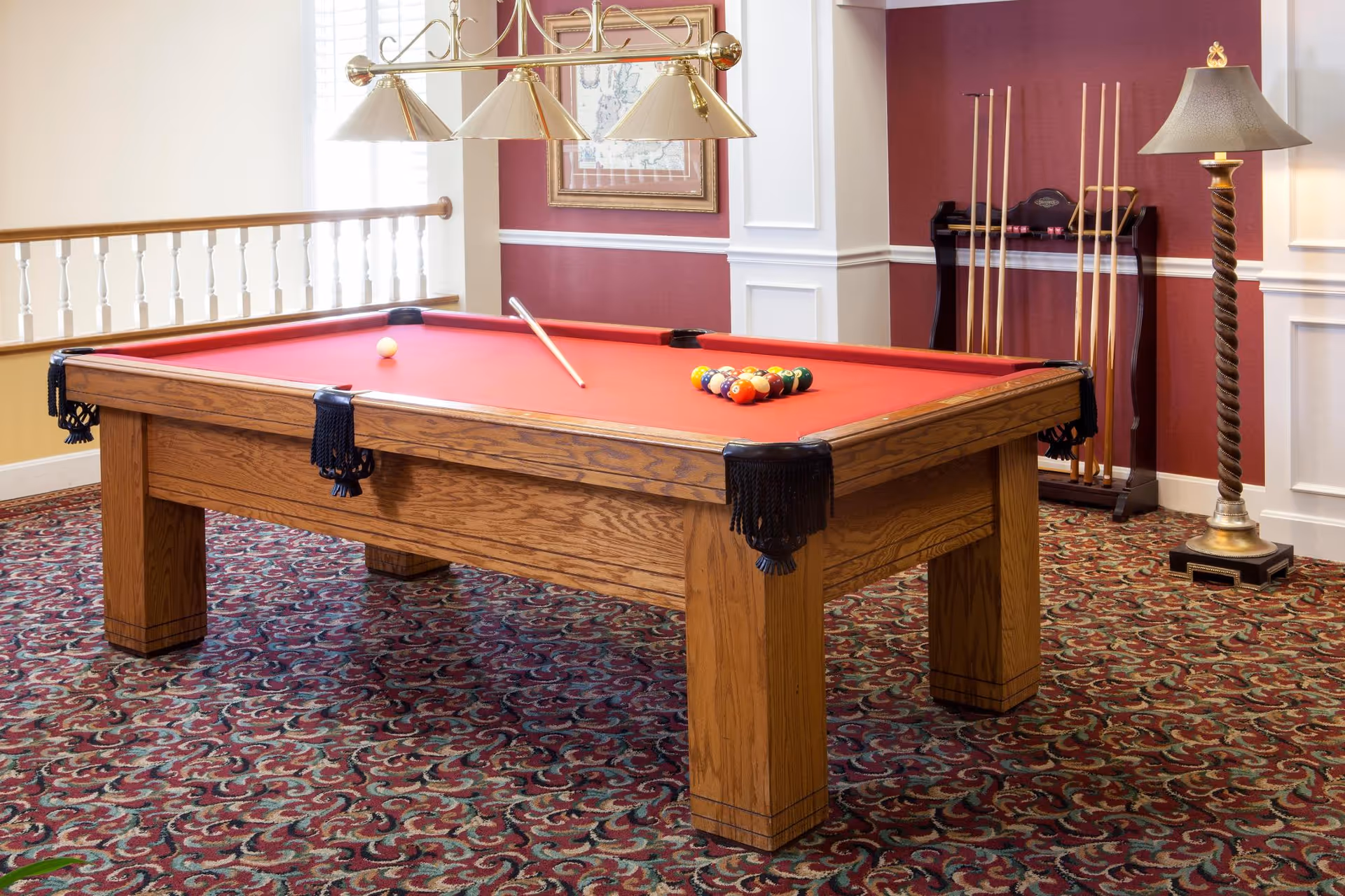 A billiards room with a wooden pool table featuring a red felt surface. The table has a cue stick resting on it and a rack of billiard balls arranged in a triangle. In the background, there is a rack holding several pool cues and a tall floor lamp with a twisted wooden stand. The room has patterned carpet, red and white walls, and a brass light fixture hanging above the table.