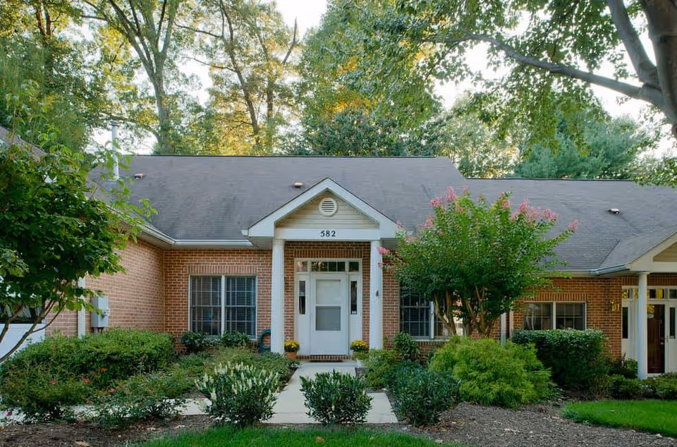 Front exterior view of a single-story brick building with a white door and two white columns at the entrance, surrounded by green shrubs, trees, and a well-maintained garden under a clear sky.