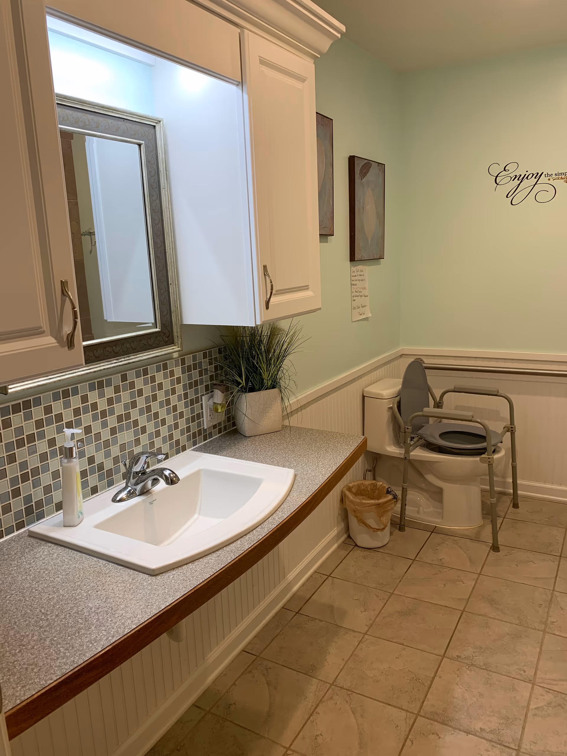 A clean bathroom with a sink and countertop on the left side, featuring a soap dispenser and a decorative plant. Above the sink are white cabinets and a mirror. On the right side is a toilet with a raised seat and safety rails, a small trash can beside it, and two framed pictures on the wall. The walls are painted light green with white wainscoting, and there is a decorative wall decal that says 'Enjoy the simple'. The floor is tiled.