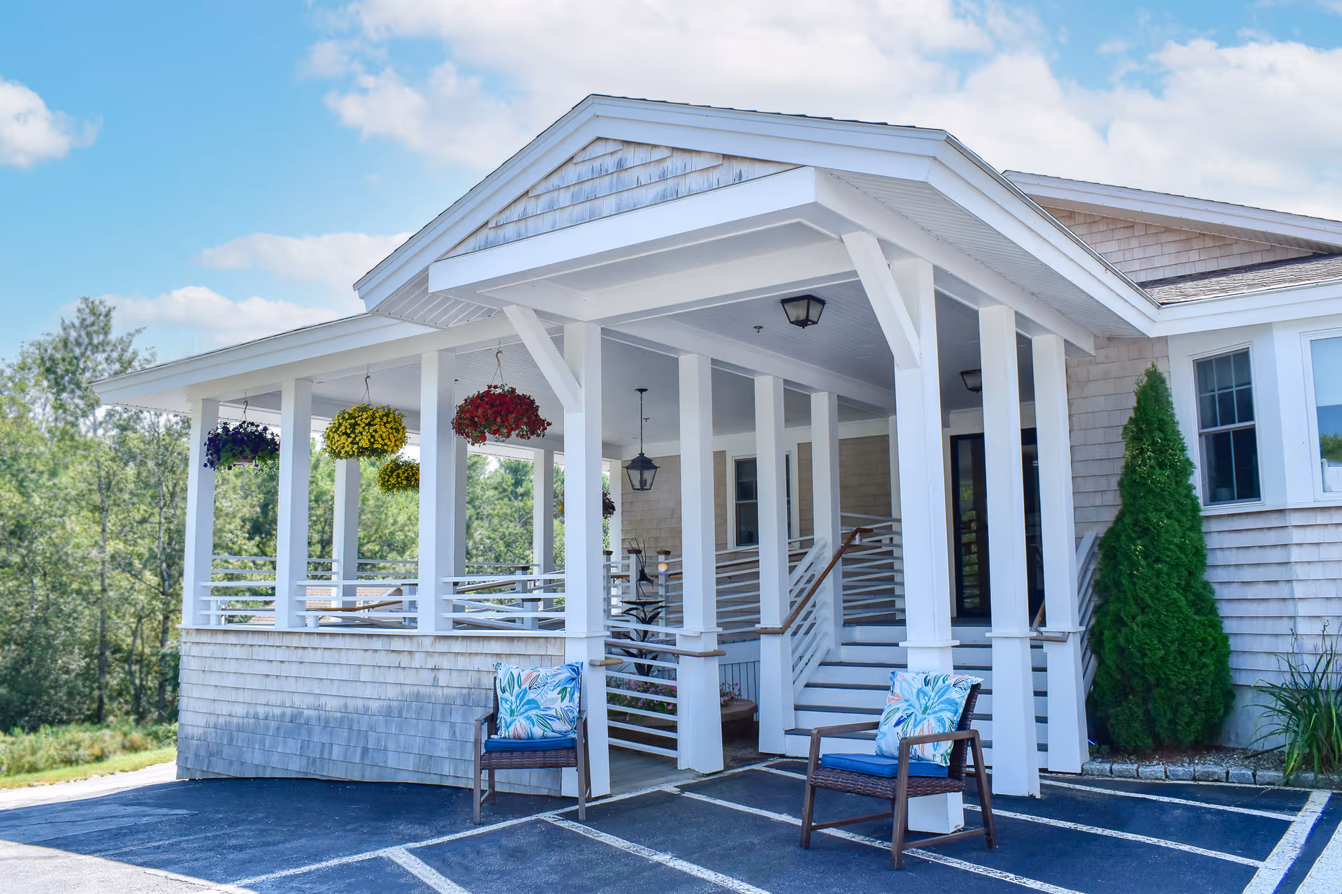 Covered front entrance of a senior living building with white columns, hanging flower baskets, and two outdoor chairs.