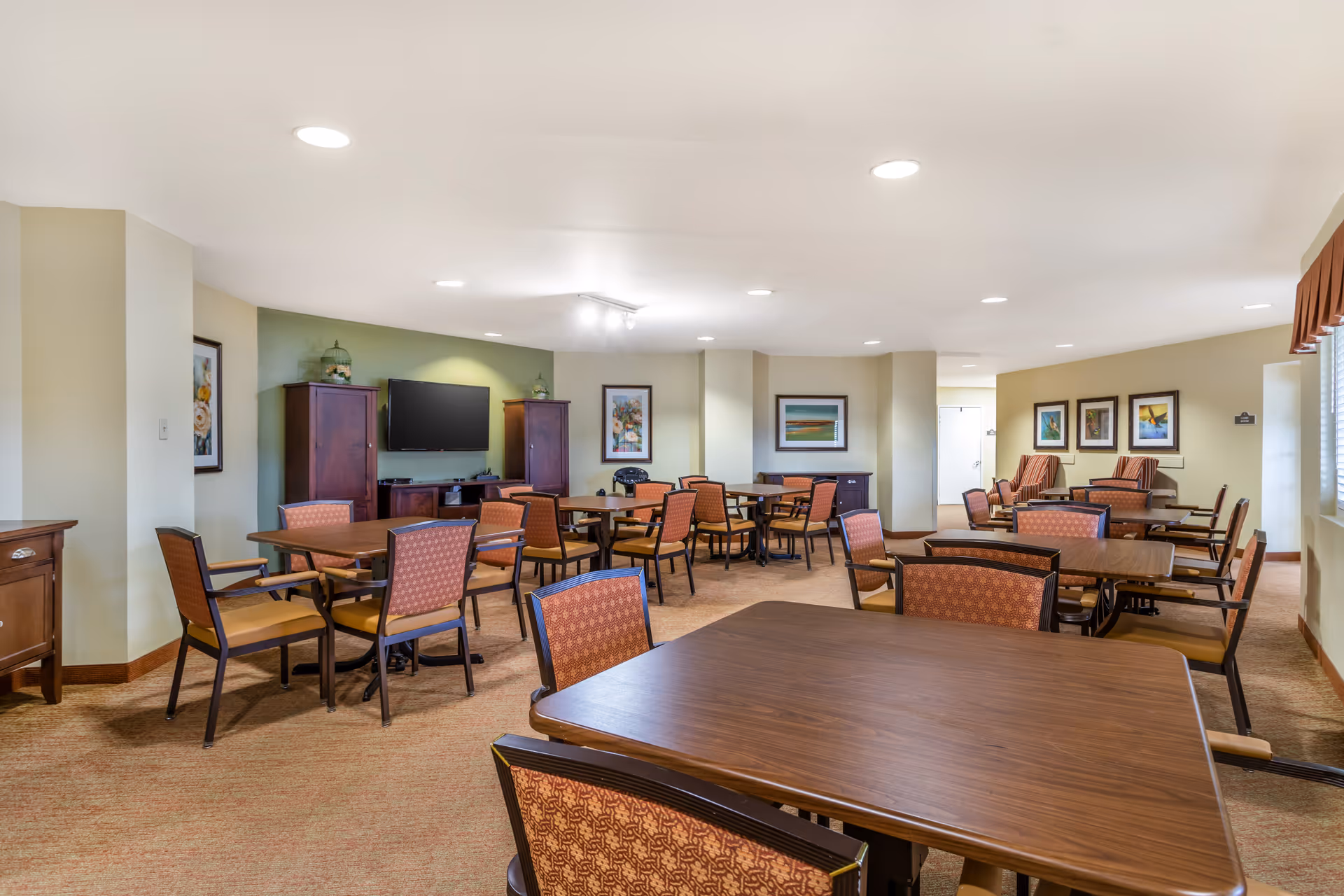 Spacious senior living common dining area with multiple tables and chairs, a television, and framed artwork on the walls.