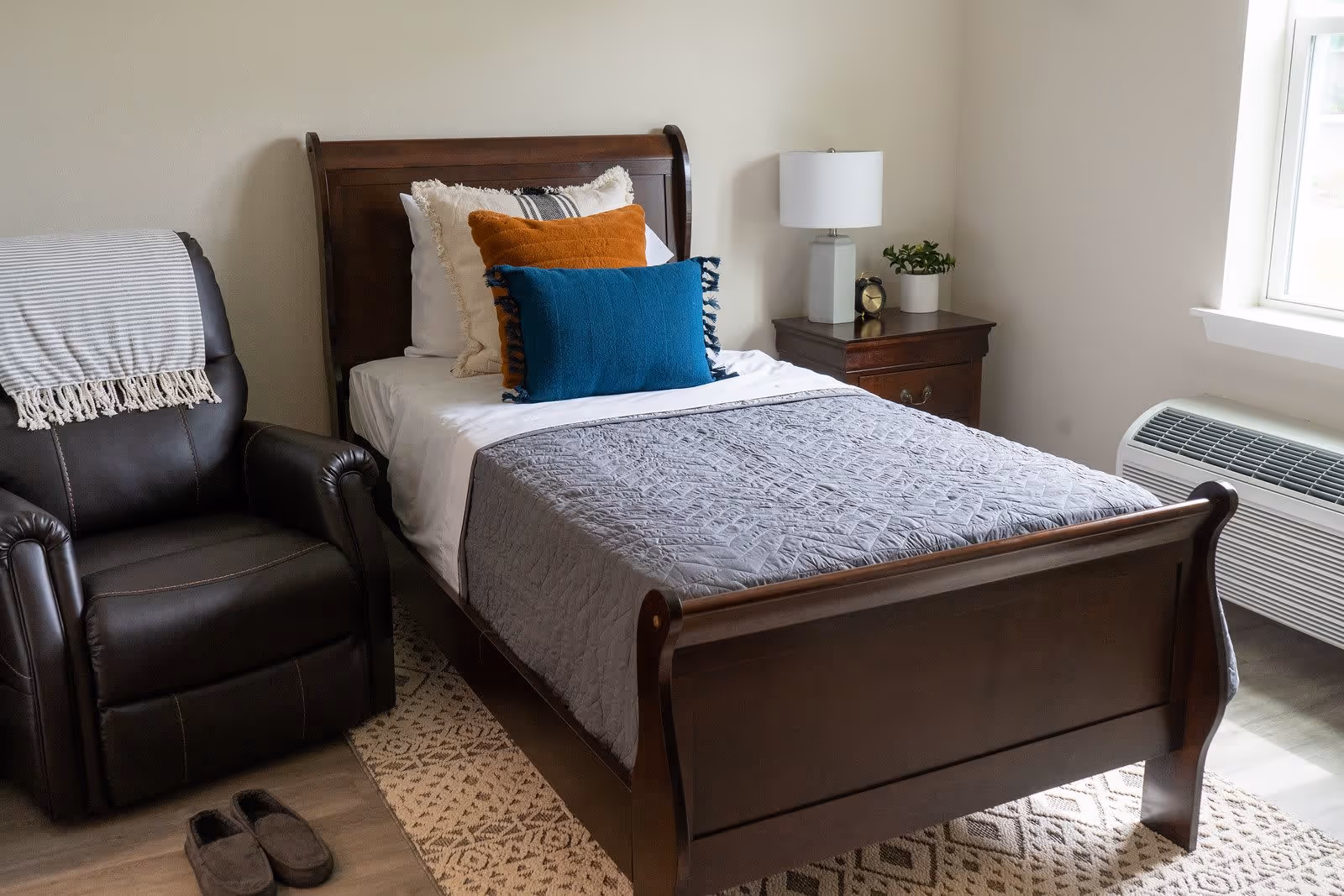 A cozy bedroom with a wooden bed frame, neatly made bed with multiple pillows in white, orange, and blue, a bedside table with a lamp, clock, and small plant, a dark brown recliner chair with a striped throw blanket, slippers on the floor, and a window letting in natural light.