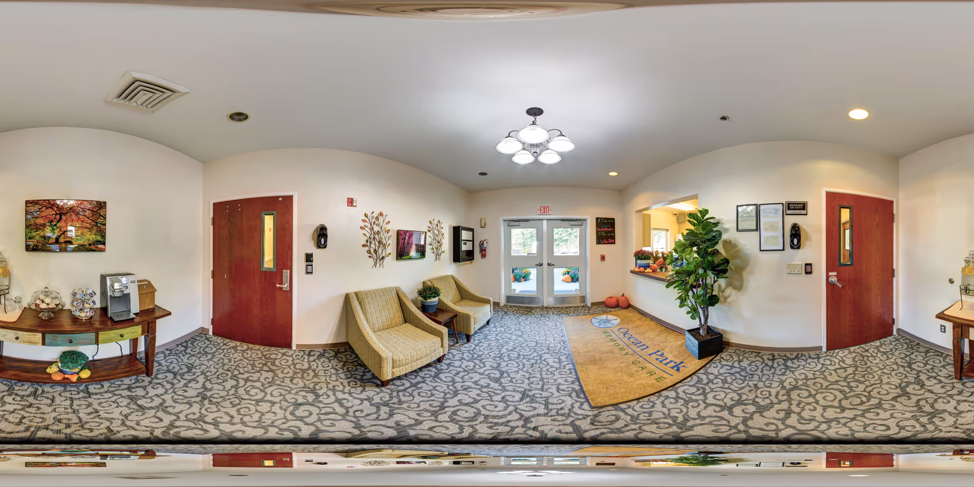 A bright and welcoming lobby area with patterned carpet, two beige armchairs with a small table and plant between them, a wooden console table with a coffee machine and decorative items, wall art including a colorful tree painting and metal leaf sculptures, a large potted plant, double glass exit doors with an exit sign above, and a mat on the floor that reads 'Ocean Park Memory Care'.