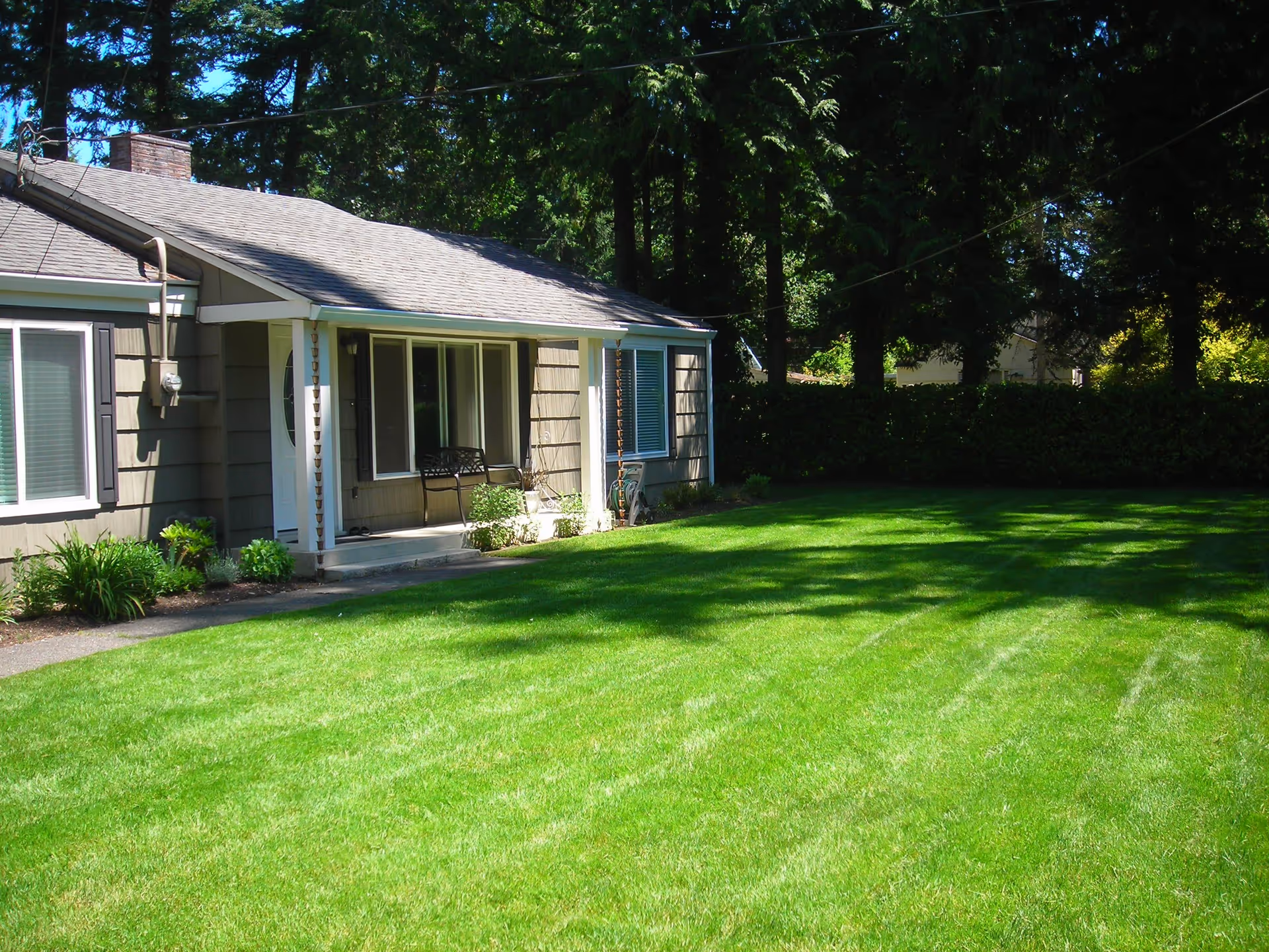 Single-story house with a gray roof and beige siding, featuring a small covered porch with a bench. The house is surrounded by a well-maintained green lawn and tall trees in the background.