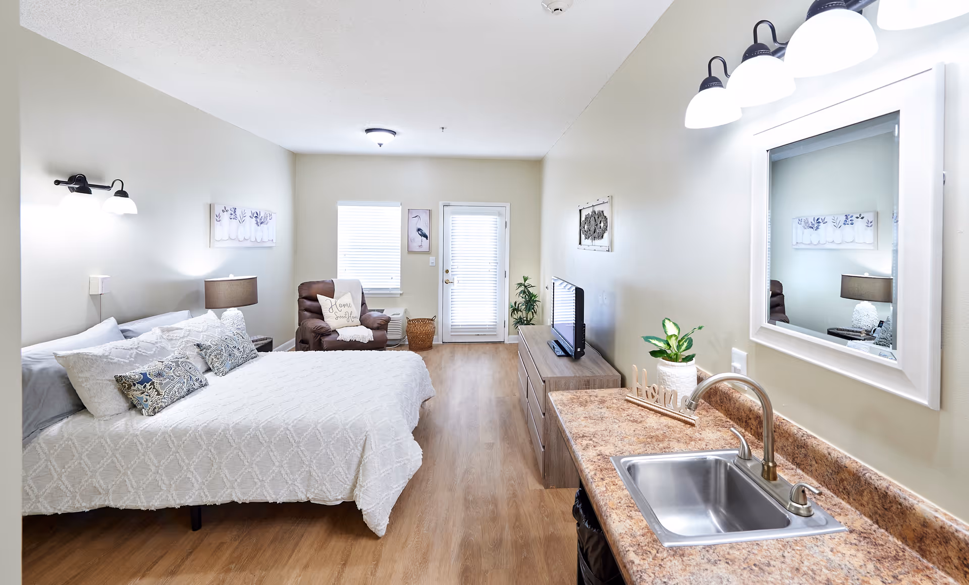Bright studio apartment interior with a bed, recliner, TV on a dresser, and a kitchenette sink and counter.