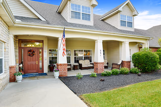 Front entrance of a senior living facility with a covered porch featuring seating, brick columns, an American flag, and a red entry door.