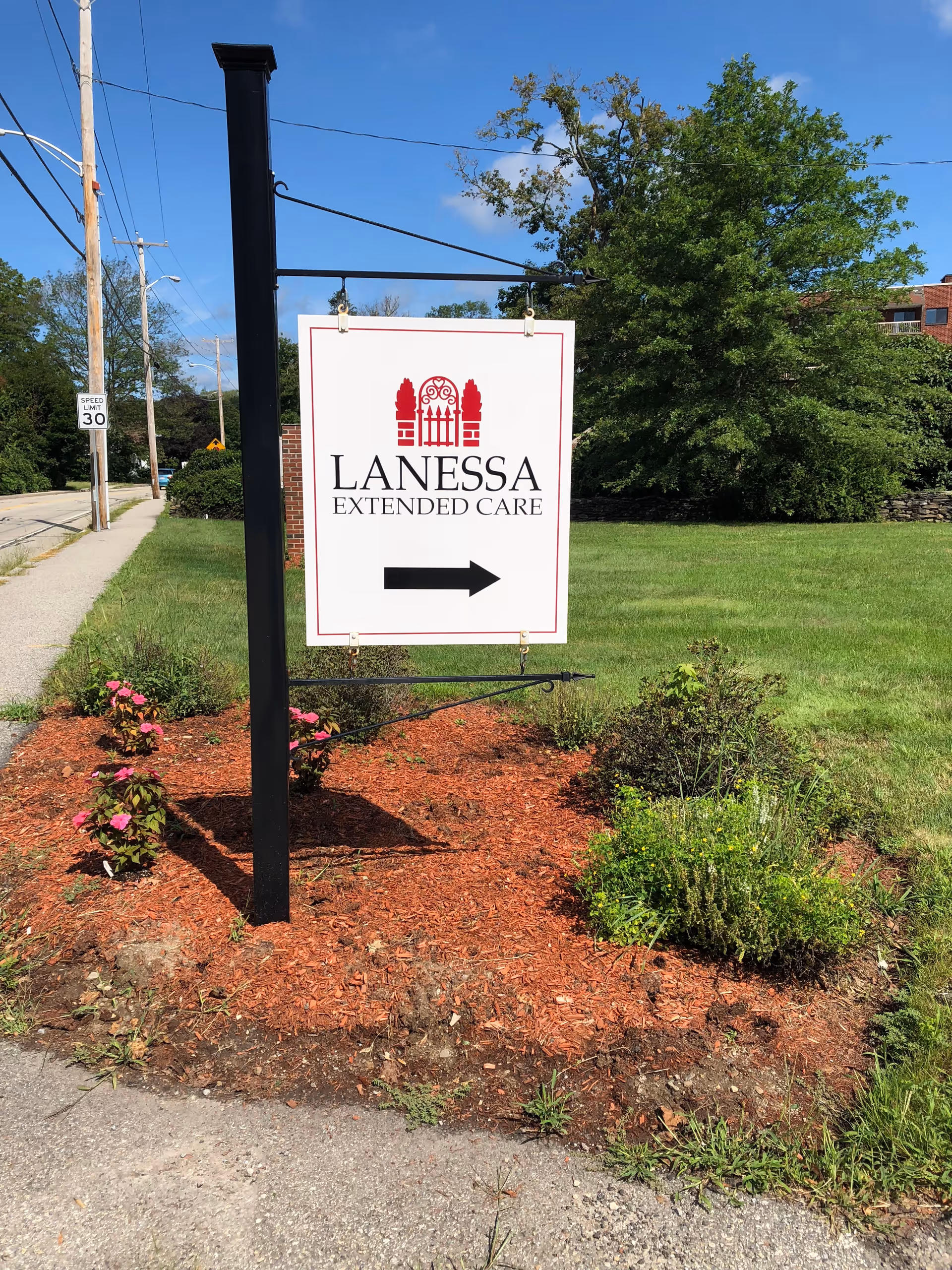 A white sign with red and black text reading 'Lanessa Extended Care' with a right-pointing arrow, mounted on a black post in a landscaped area with mulch, flowers, and greenery. The background shows a sidewalk, street, utility poles, and trees under a clear blue sky.