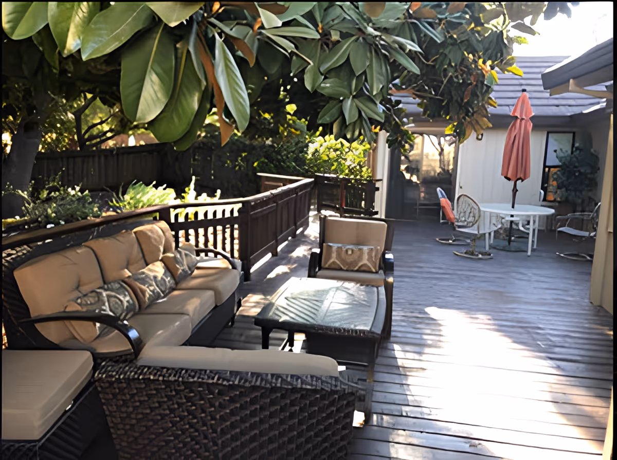 Outdoor patio area with cushioned wicker seating, a glass-top coffee table, and a round table with chairs and a closed pink umbrella. The patio is surrounded by greenery and shaded by large leafy branches.