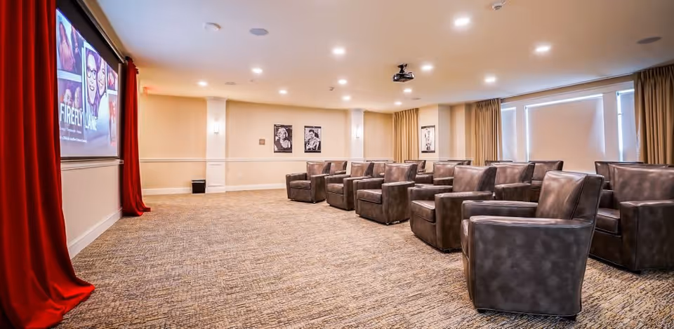 A cozy movie theater room with multiple rows of comfortable brown leather armchairs facing a large screen displaying the movie Firefly Lane. The room has beige walls, carpeted floor, red curtains framing the screen, and framed black and white photos on the walls. Ceiling lights illuminate the space.