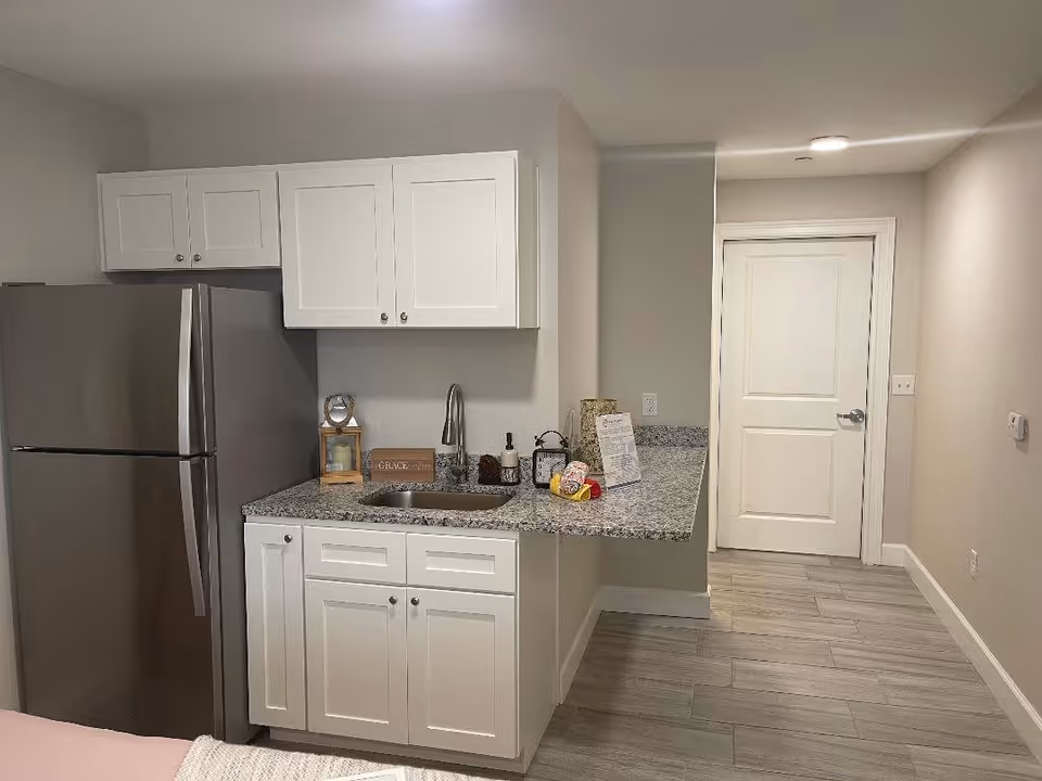 A small kitchen area with white cabinets, a stainless steel refrigerator, a granite countertop with a sink, and various decorative items including a candle, a small clock, and a sign that says 'Grace Manor'. The floor is tiled with light gray tiles, and there is a white door at the end of the hallway.