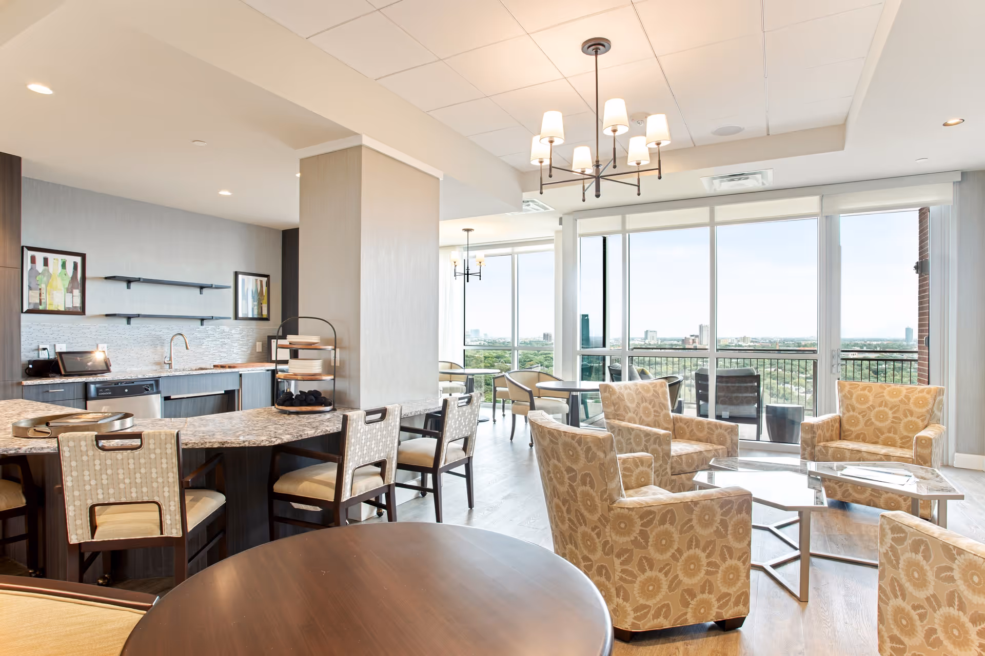 Bright and modern common area in a senior living facility featuring a kitchen with granite countertops and bar stools, a seating area with patterned armchairs around a glass coffee table, large windows offering a city view, and a balcony with outdoor chairs.