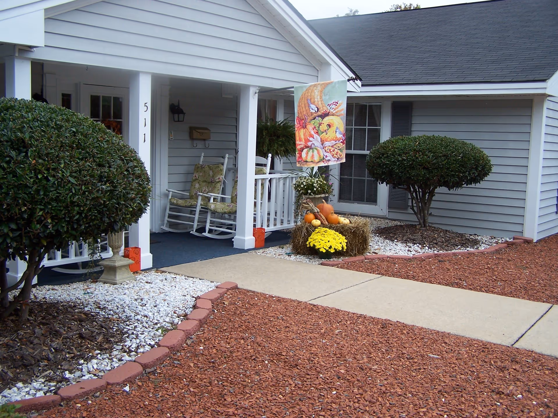 Front porch area of a retirement facility with two cushioned rocking chairs, a mailbox, and a decorative fall-themed flag with pumpkins and gourds. The porch is surrounded by neatly trimmed bushes and landscaping with white rocks and red mulch.