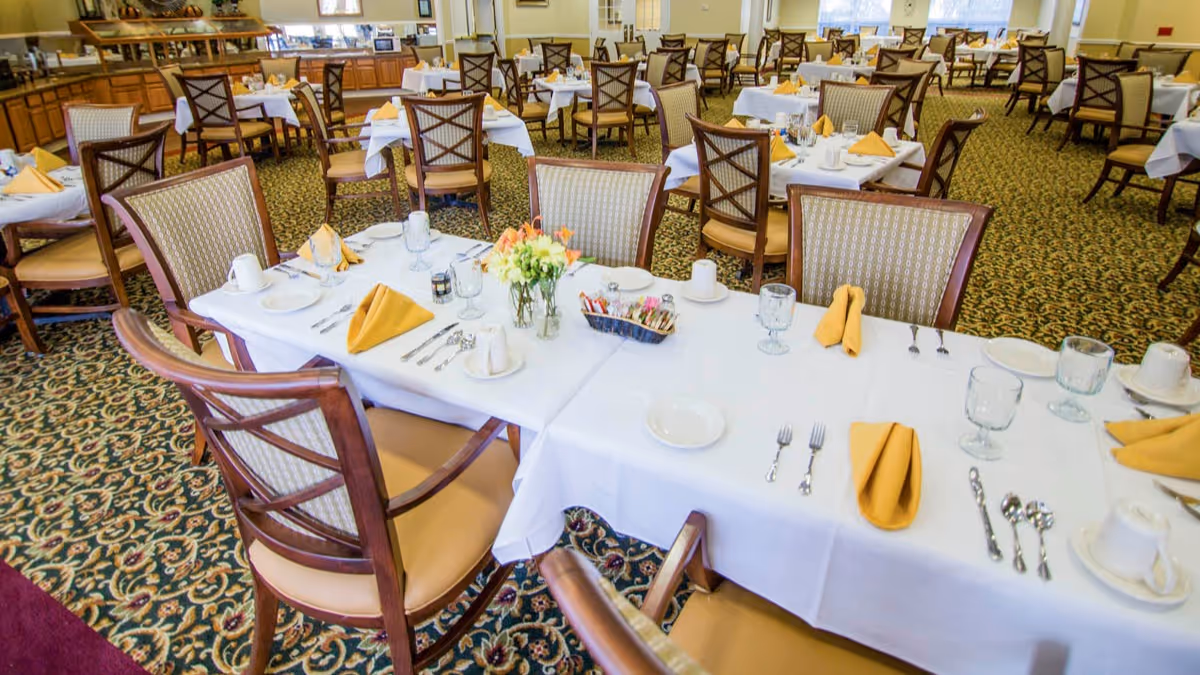 A spacious dining room with multiple tables covered in white tablecloths, set with plates, glasses, silverware, and yellow folded napkins. The room has patterned carpet flooring and wooden chairs with cushioned seats and backs. A buffet area with cabinets and food trays is visible in the background.