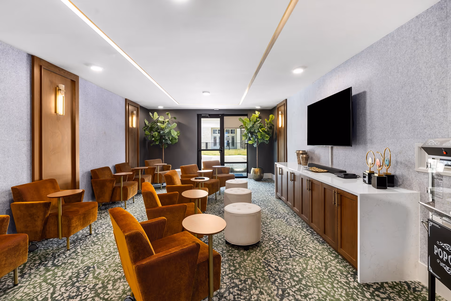 A modern lounge area with multiple orange upholstered armchairs each paired with a small round wooden table. The room features a patterned carpet, light gray walls with wooden panel accents, and two large potted plants near a glass door leading outside. On the right wall, there is a mounted flat-screen TV above a long white countertop with wooden cabinets underneath. A popcorn machine is visible on the far right side.