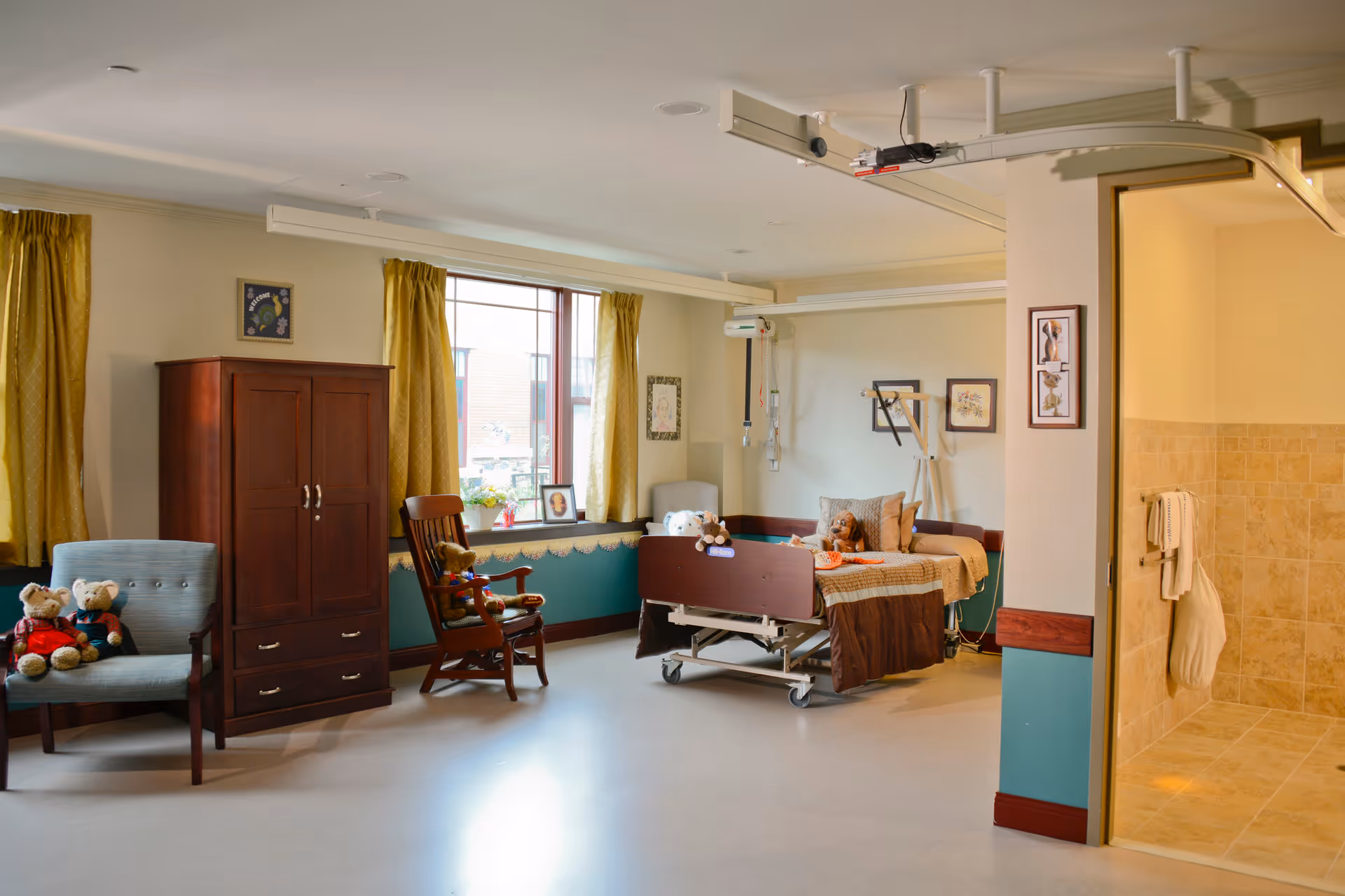 A well-lit senior living facility room with a hospital bed, a wooden wardrobe, a rocking chair, and an armchair. The bed has pillows and stuffed animals on it. There are yellow curtains on the windows, framed pictures on the walls, and a tiled bathroom with towels visible through an open doorway.