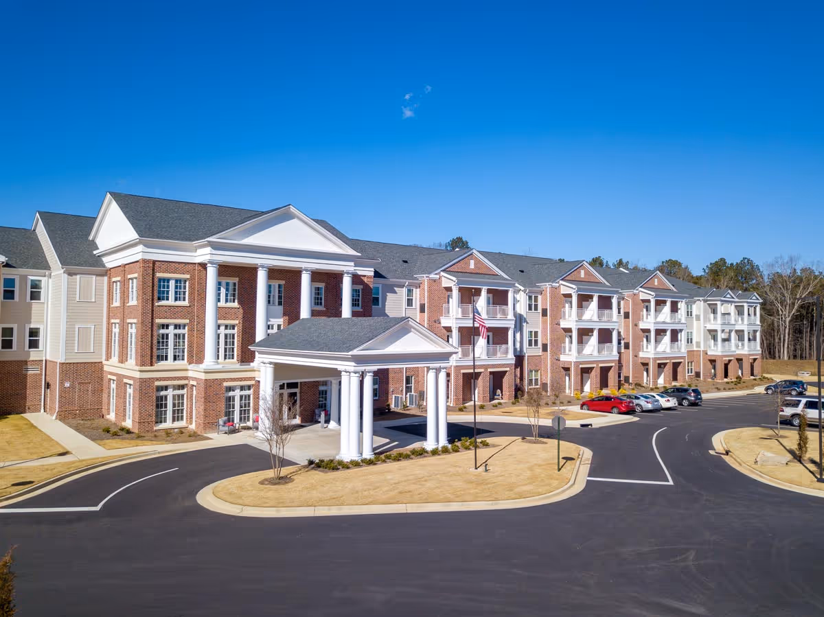 Exterior view of a large senior living facility building with red brick and white columns, multiple balconies, a covered entrance, a parking lot with cars, and a clear blue sky.