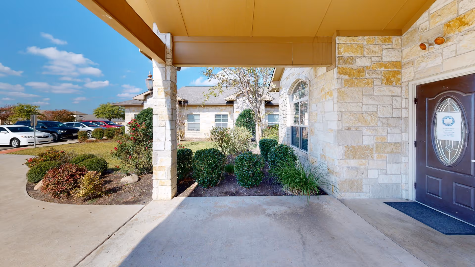 Covered stone entryway and driveway of a senior living building with landscaped shrubs, parked cars in the background, and a front door with a posted notice.