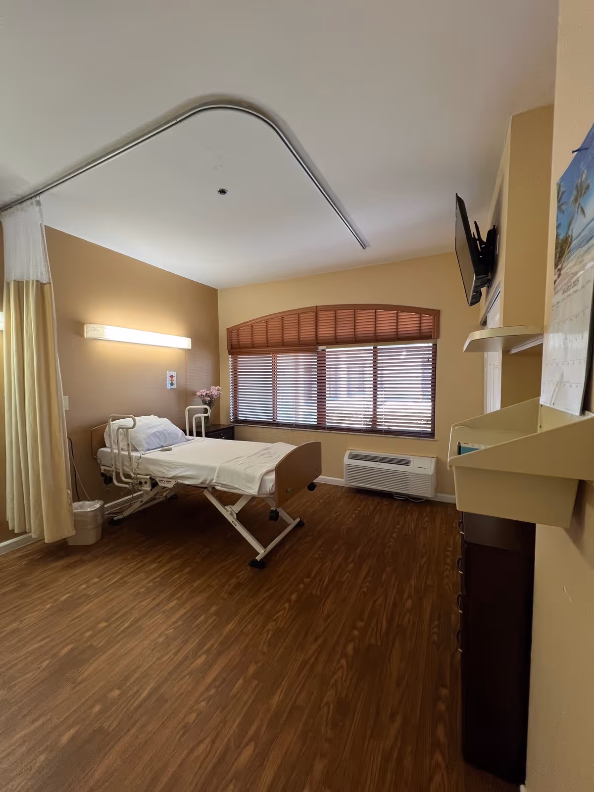 A clean senior facility bedroom with an adjustable hospital bed, window blinds, a wall-mounted TV and wood-look flooring.