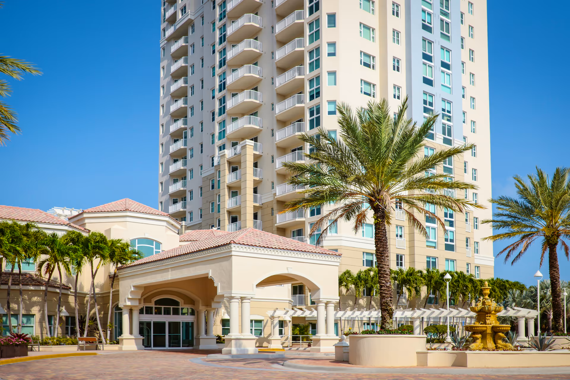 Exterior view of a multi-story beige residential building with balconies, an arched entrance, palm trees, and a fountain.