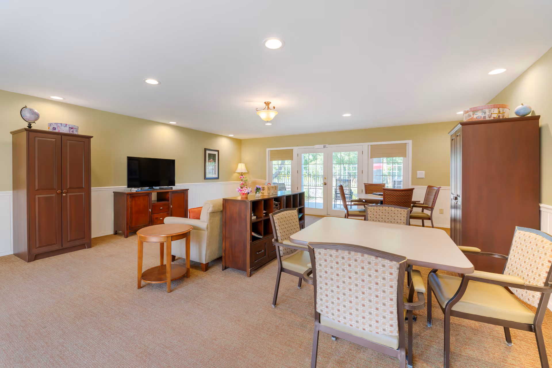 A bright and spacious common area in a senior living facility featuring a seating area with a beige sofa, wooden cabinets, a flat-screen TV, and multiple tables with chairs. The room has beige walls with white wainscoting, carpeted floors, recessed lighting, and large windows with glass doors leading outside.