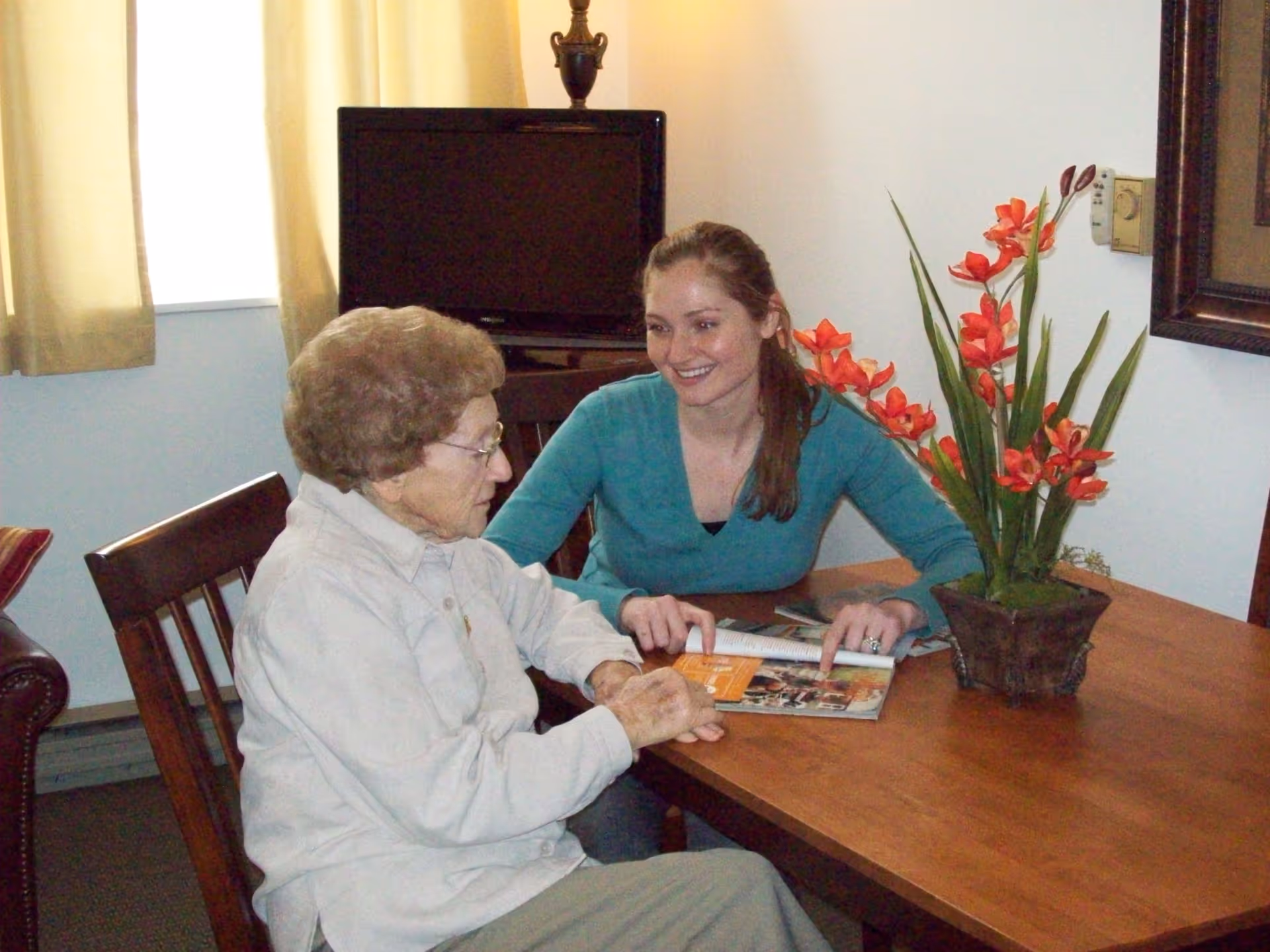 An elderly woman and a younger woman sitting at a wooden table in a room. The younger woman is smiling and pointing at a magazine on the table. There is a potted plant with red flowers on the table and a television in the background near a window with beige curtains.
