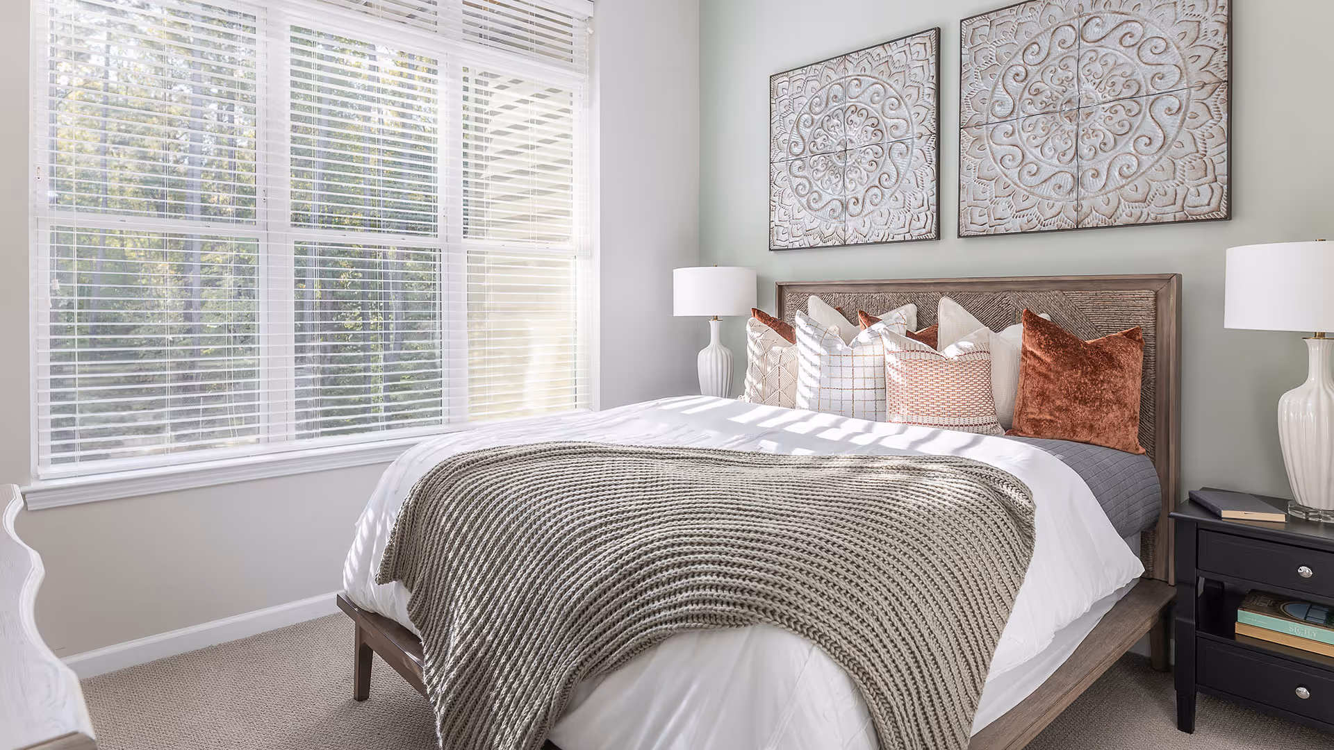 A bright bedroom with a large window covered by white blinds letting in natural light. The bed has a wooden frame and is made with white bedding, a gray knitted throw blanket, and multiple decorative pillows in white, beige, and rust colors. Two white table lamps sit on black nightstands on either side of the bed. Above the bed are two square decorative wall art pieces with intricate patterns.