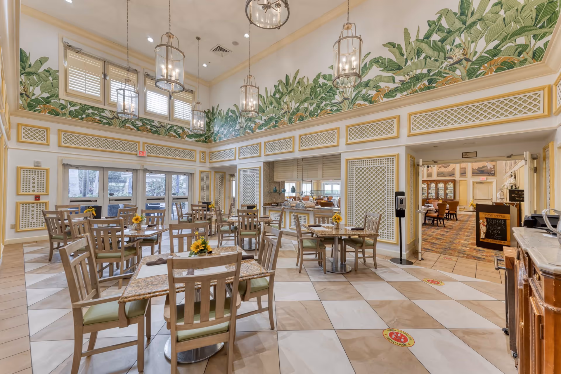 Bright dining room with multiple wooden tables and chairs, hanging lanterns, a tropical leaf mural near the high ceiling, and a serving area.