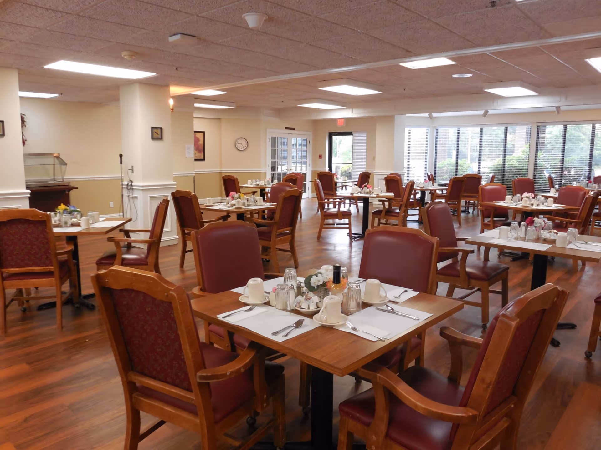 Spacious dining room with multiple set tables and wooden chairs arranged for a meal in a senior living facility.