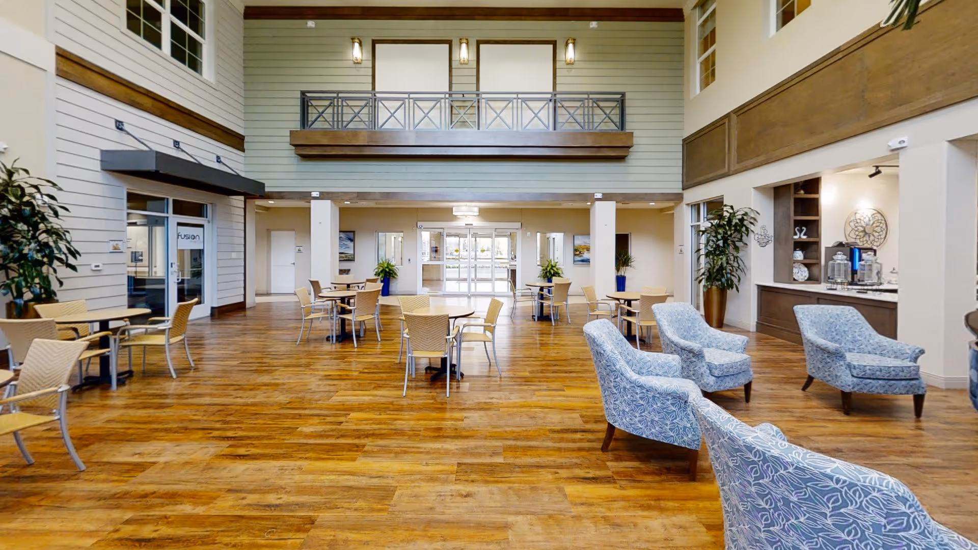 Spacious and well-lit common area with wooden flooring, several round tables with beige chairs, and a seating area with blue patterned armchairs. The room has high ceilings, large windows, potted plants, and a balcony railing above the entrance doors.