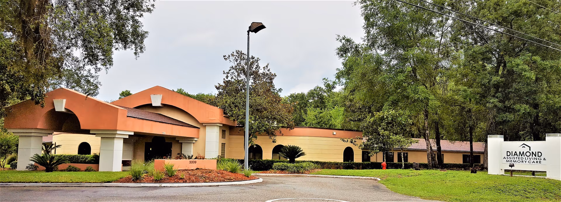 Exterior view of Diamond Assisted Living & Memory Care facility showing a single-story building with a covered entrance, surrounded by trees and greenery. A sign with the facility name is visible on the right side near the driveway.