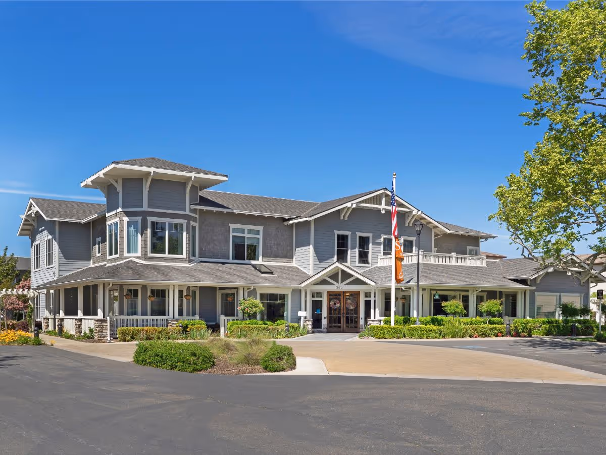 Two-story gray senior living facility building with a covered main entrance, flagpoles and landscaped grounds under a clear blue sky.