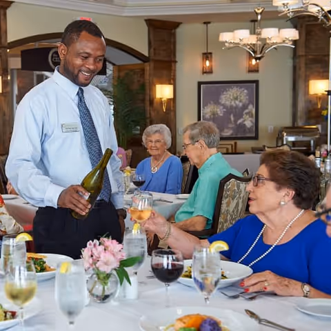 A smiling waiter in a light blue shirt and tie pours wine for an elderly woman seated at a dining table. Other elderly people are seated around the table enjoying their meals in a warmly lit dining room with framed floral artwork on the wall.