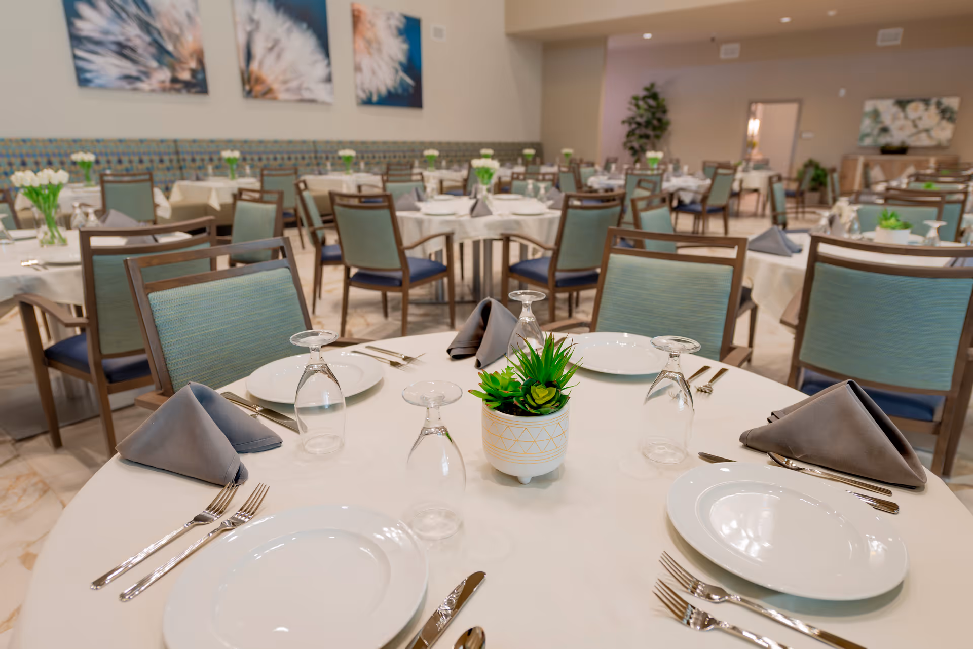 A bright assisted-living dining room with round tables set with plates, napkins, glassware, and small potted centerpieces.