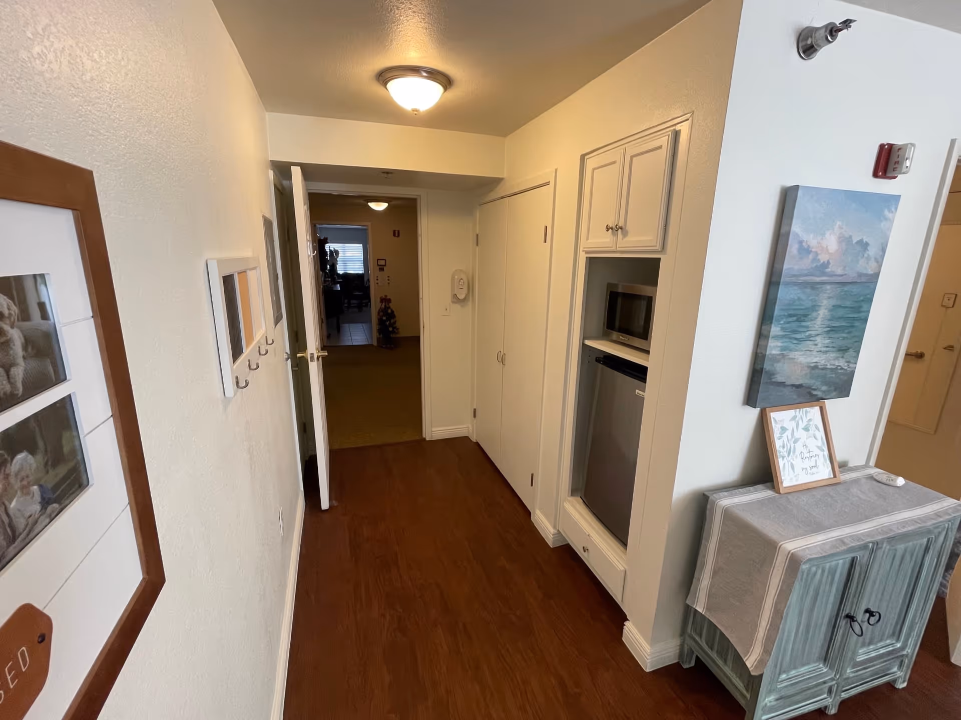 A hallway in a senior living facility with wooden flooring and white walls. On the left wall, there are framed pictures and a small coat rack with hooks. On the right side, there is a small cabinet with a tablecloth, a framed picture, and a painting of a seascape above it. Further down the hallway, there is a built-in microwave and mini refrigerator. The hallway leads to another room with carpeted flooring and a partially open door.