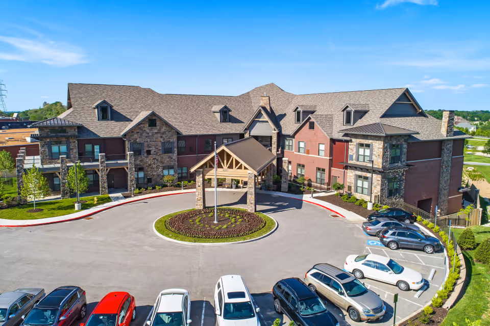 Front exterior view of a large, multi-story senior living facility with stone and brick facade, a covered entrance with wooden beams, a circular driveway with a landscaped center island, and a parking lot with several cars parked. The sky is clear and blue.