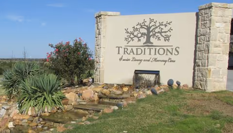 Stone entrance sign for Traditions Senior Living and Memory Care with a tree logo, small rock waterfall fountain, and surrounding landscaping under a blue sky.