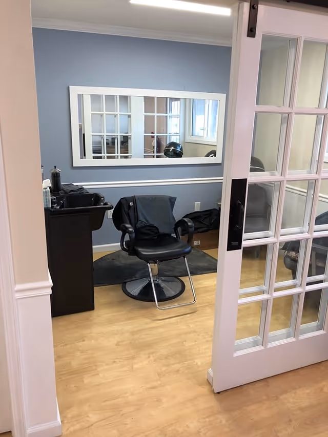 Interior view of a small salon or grooming area in a senior living facility. The room has light wood flooring, a blue-gray wall with white trim, and a large rectangular mirror with a white frame. There is a black salon chair in the center with a black cape draped over it, and a black sink station to the left. A sliding glass door with white framing is partially open, revealing the room.