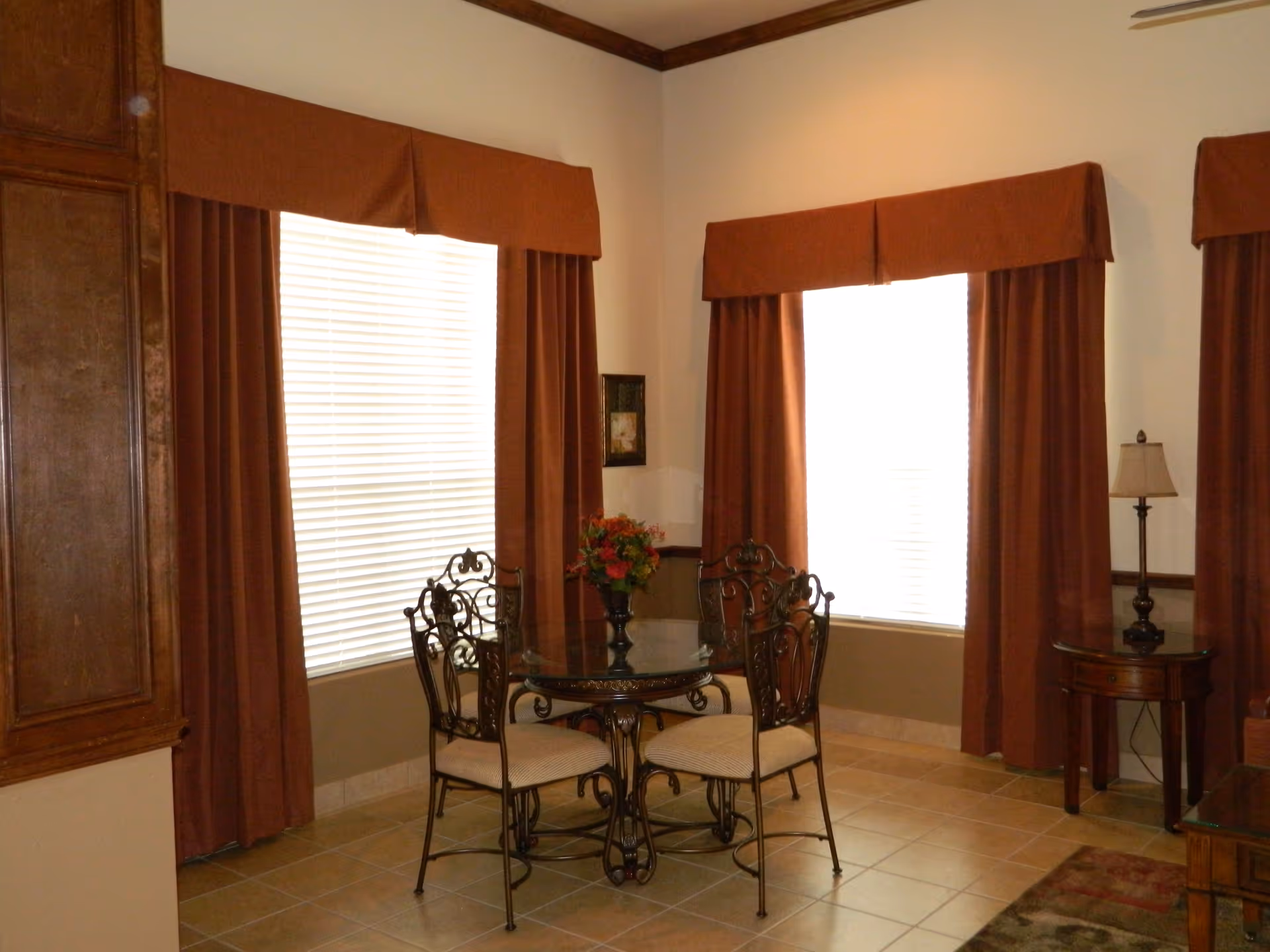 A small dining area with a round glass table and four ornate metal chairs with cushioned seats. The room has large windows covered with brown curtains and valances. There is a small side table with a lamp in the corner and a flower arrangement on the dining table. The floor is tiled and the walls are painted white with a wooden trim near the ceiling.
