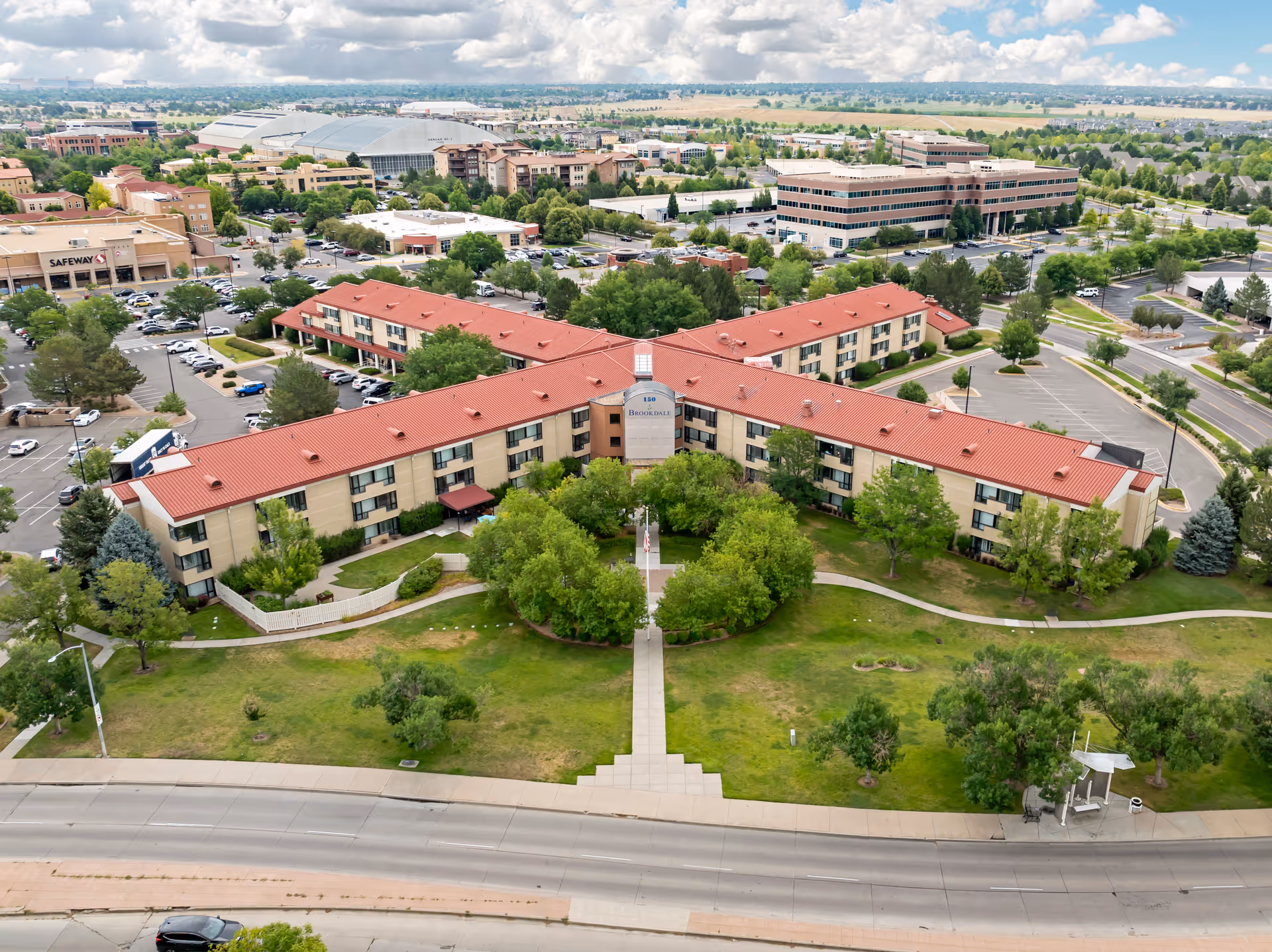 Aerial view of Brookdale Lowry, a senior living facility with a red roof and beige walls, surrounded by green lawns and trees. The building is shaped like a wide V with a central entrance and pathways leading to it. Nearby parking lots, roads, and other buildings are visible under a partly cloudy sky.