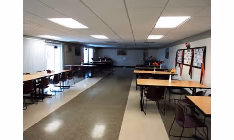 A large room with multiple tables and chairs arranged in rows, fluorescent ceiling lights, and bulletin boards on the walls. The room appears to be a communal dining or meeting area with windows on one side letting in natural light.