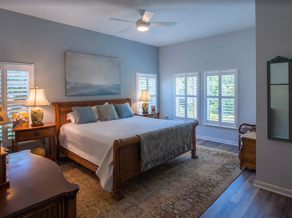 A well-lit bedroom with a large wooden bed featuring white bedding and multiple pillows. There are two bedside tables with lamps on each side of the bed. The room has light blue walls, three windows with white shutters, a large area rug with intricate patterns, and a ceiling fan with a light fixture. A mirror is mounted on the wall to the right.