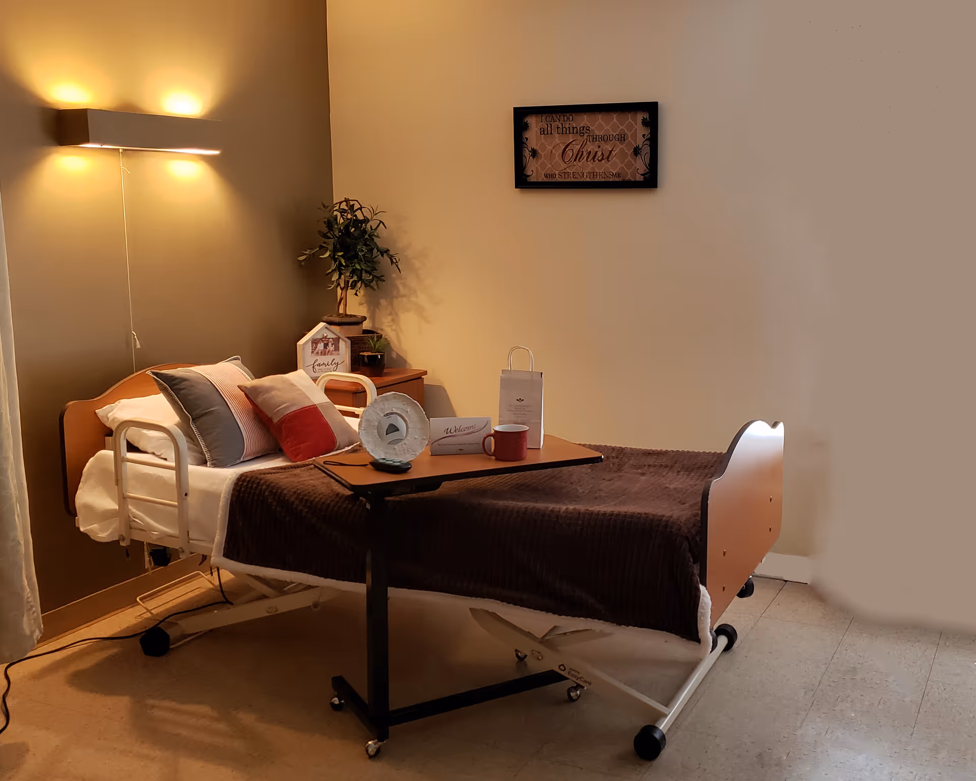 A senior living facility bedroom with a hospital-style bed covered with a brown blanket and several pillows. A wooden bedside table holds a small plant and decorative items. A wall-mounted light fixture illuminates the room softly. A framed inspirational sign hangs on the wall above the bed. A rolling overbed table holds a red mug, a welcome card, and a small gift bag.