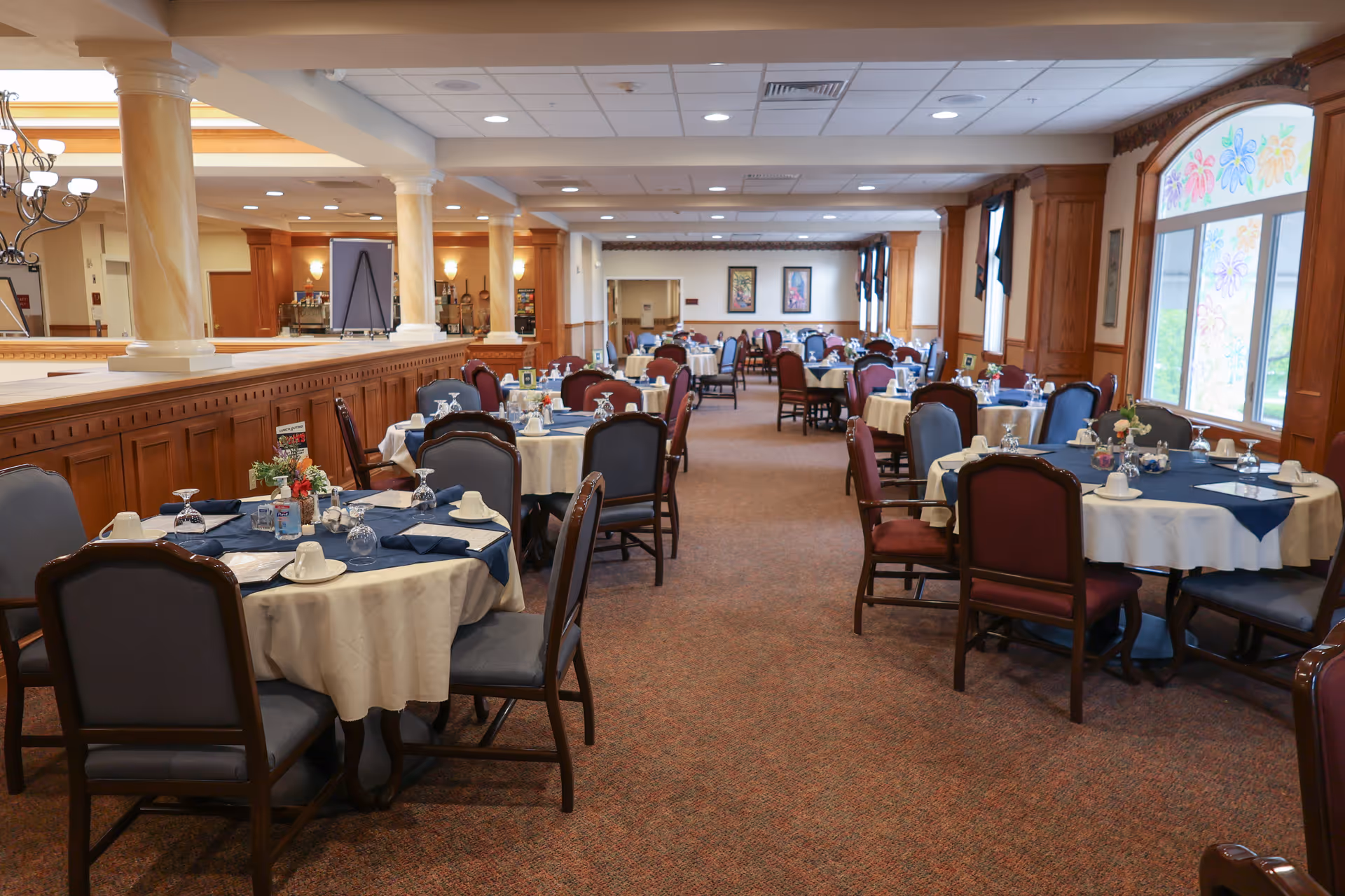 A spacious dining room with multiple round tables covered with white and blue tablecloths, set with cups, glasses, and napkins. The room features wooden paneling, large windows with floral decorations, and comfortable chairs arranged around the tables. The ceiling has recessed lighting and decorative columns separate the dining area from an adjacent space.