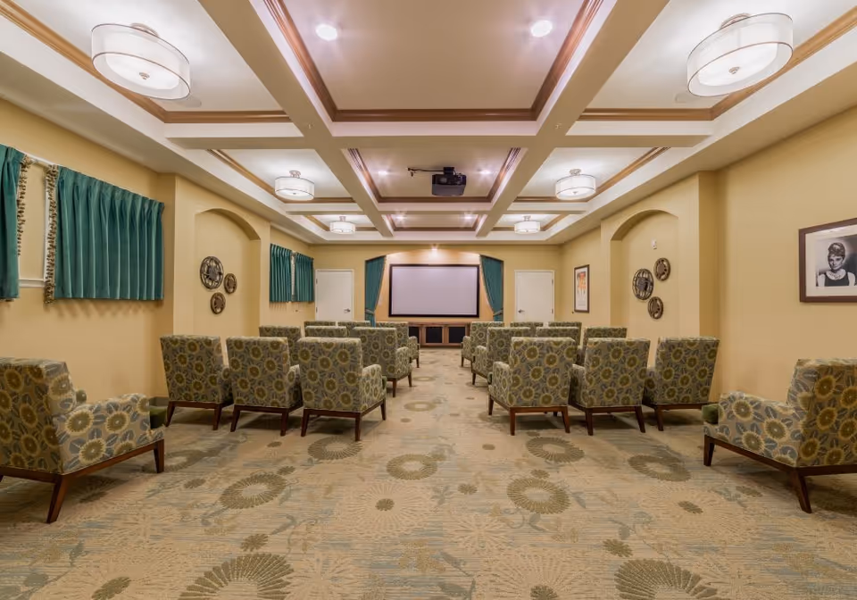 Carpeted community media room with patterned armchairs arranged in rows facing a projection screen.