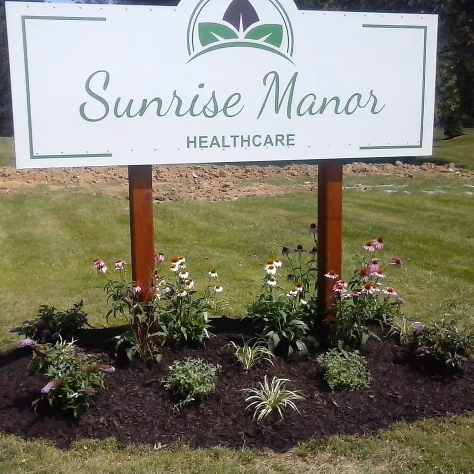 A white sign with green text that reads 'Sunrise Manor Healthcare' mounted on two wooden posts, surrounded by a flower bed with various blooming flowers and green grass in the background.