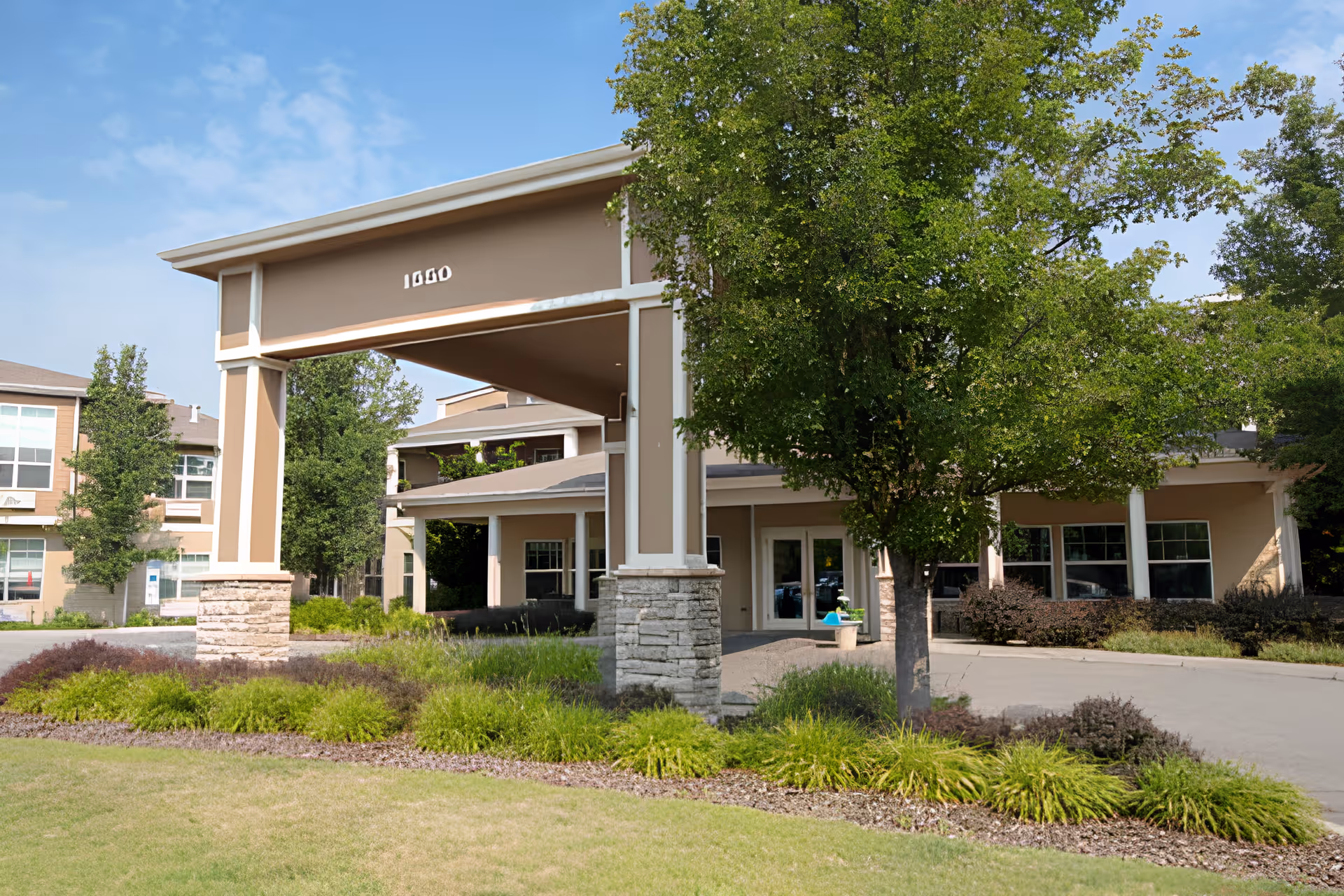 Entrance of Wenatchee Senior Living by Cogir facility showing a covered driveway with pillars, surrounded by green landscaping and trees under a blue sky.