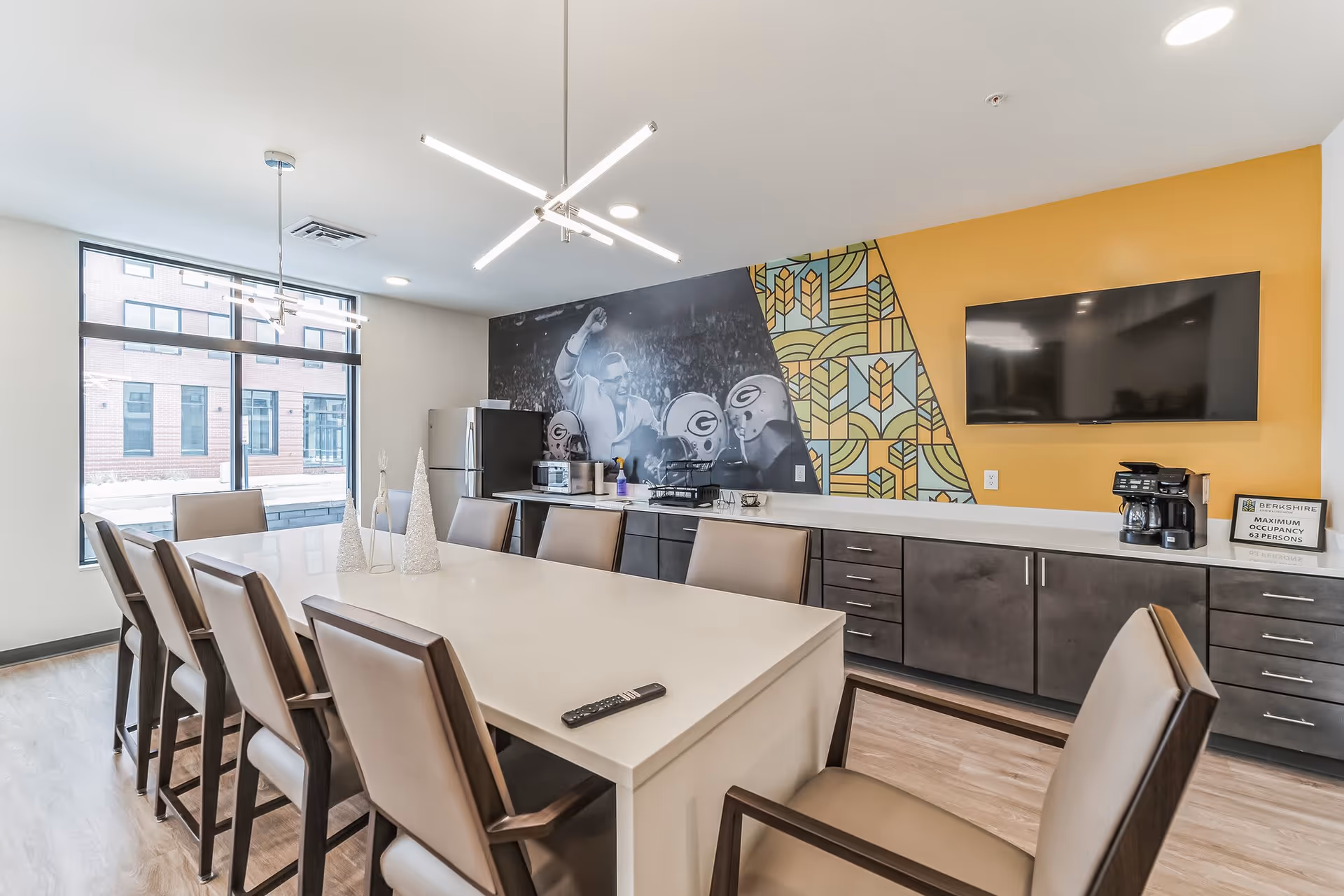 A modern dining room with a long white table surrounded by beige cushioned chairs. The room features a large window letting in natural light, a refrigerator, microwave, coffee maker, and a flat-screen TV mounted on a yellow accent wall. A mural with a sports theme and geometric patterns decorates one wall.