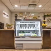 Interior view of a senior living facility's refreshment area with a glass display cooler containing bottled water and green apples. The counter behind the cooler has decorative plants and flowers, with a sign on the wall reading 'Discovery Village At Stuart'.